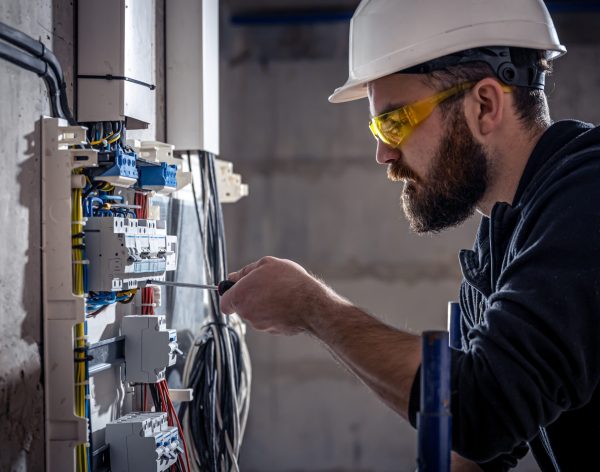 A male electrician works in a switchboard with an electrical connecting cable, connects the equipment with tools.