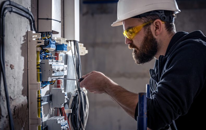 A male electrician works in a switchboard with an electrical connecting cable, connects the equipment with tools.